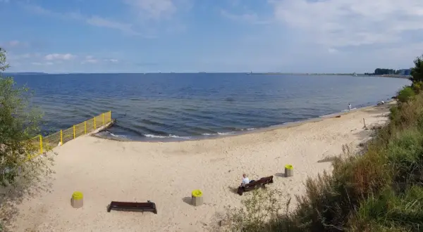 Plaża dla psów z łagodnym zejściem do morza. Na piasku ławki i kosze. W oddali widać falochron.