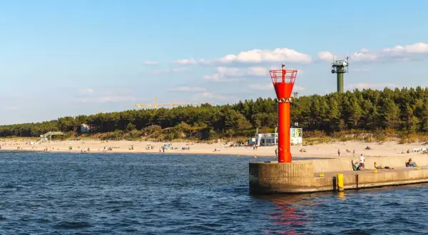 Mrzeżyno atrakcje: plaża, morze i charakterystyczna czerwona wieża nawigacyjna na falochronie. W tle las i zielona wieża.