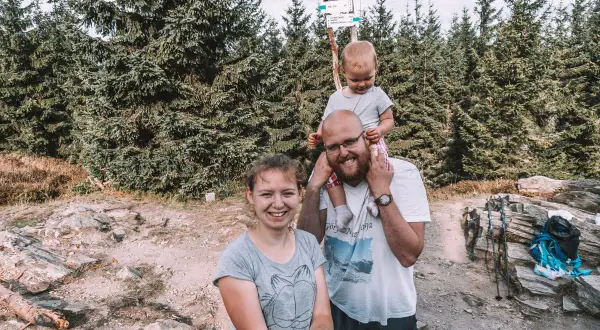 Rodzina na szlaku. Dziecko na barana, rodzice uśmiechnięci. W tle las i drogowskaz z napisem "Kowadło szczyt".