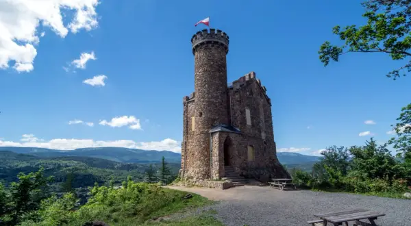 Kamienny zamek księcia Henryka na trasie widokowej, z polską flagą na wieży, otoczony zielonymi wzgórzami.