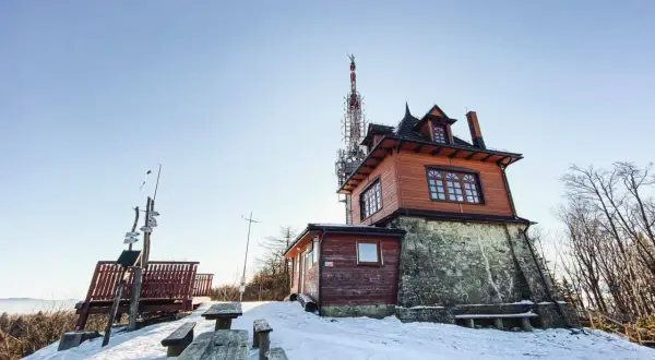 Zimowy krajobraz na Luboniu Wielkim. Drewniany budynek schroniska, wieża telekomunikacyjna i ławki w śniegu.