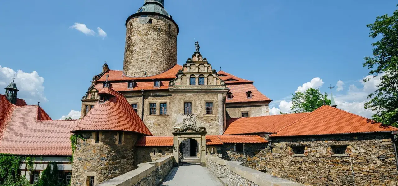 Zamek Czocha, malownicza trasa turystyczna. Kamienne mury, czerwone dachy i wieża zamkowa pod błękitnym niebem.