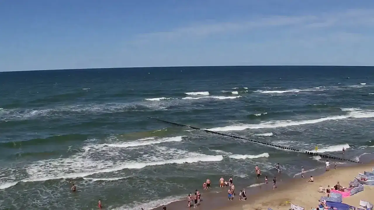 Słoneczny dzień na plaży w Rewalu. Ludzie kąpią się w morzu, a fale łagodnie rozbijają się o brzeg. To jedne z wielu rewal atrakcje.