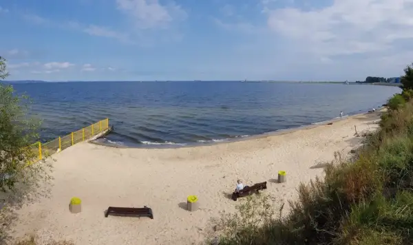 Plaża dla psów z łagodnym zejściem do morza. Na piasku ławki i kosze. W oddali widać falochron.