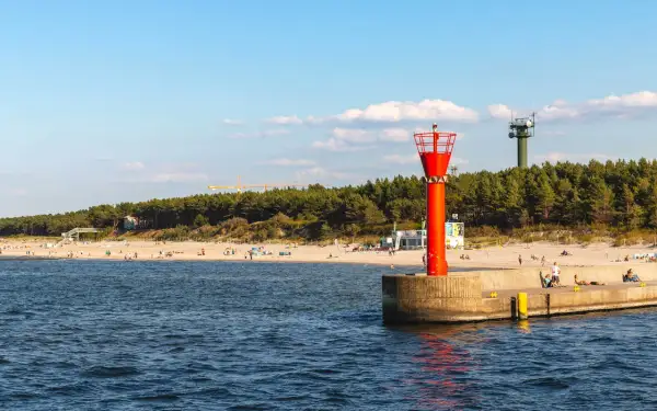 Mrzeżyno atrakcje: plaża, morze i charakterystyczna czerwona wieża nawigacyjna na falochronie. W tle las i zielona wieża.