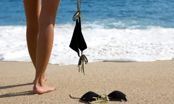 Nogi na piaszczystej plaży, obok czarny biustonosz. W tle błękitne morze i biała piana fal. Idealne miejsce na plażę nudystów.