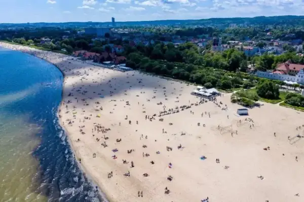 Idealny dzień, by wsiąść w pociąg i pojechać nad morze. Szeroka, piaszczysta plaża pełna ludzi, z błękitną wodą i zielonym miastem w tle.